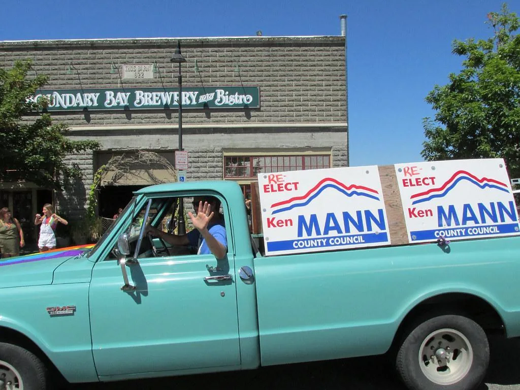 A turquise truck drives through town sporting political campaign signs. The driver waves to the people on the street.